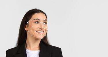 A woman stands facing slightly to the side with a smile, as facial recognition markers appear on her face. The background is plain, emphasizing the focus on technology in human expressions. photo