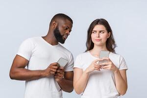 Curious Black Man Spying And Peeking At Smartphone Of His Girlfriend While They Standing Together Over Light Background In Studio, Empty Space photo