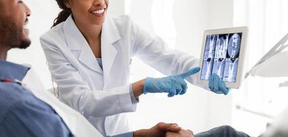 Smiling african female dentist showing young man patient teeth xray, using digital tablet photo