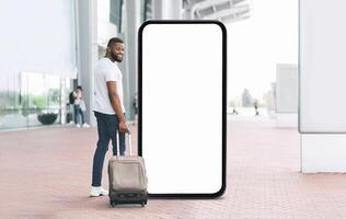 A young black man smiles at the camera while standing beside a large blank screen. He has a suitcase by his side, suggesting he is ready for travel at a busy transportation location. photo