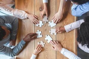 Collaboration concept, top view of creative multiracial team sitting around table and putting together puzzle pieces, cropped photo