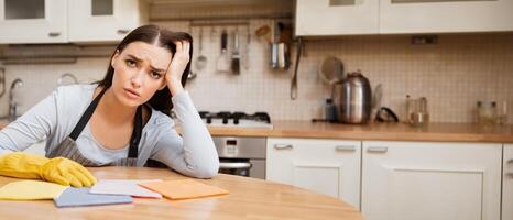 Kitchen Chores Concept. Frustrated maid sitting at dining table with cleaning cloth, resting head on hand, looking at camera photo