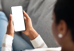 Unrecognizable Black Woman Holding Smartphone With White Screen At Home, Over Shoulder View Of African American Lady Using Blank Mobile Phone While Relaxing On Couch, Mockup Image With Copy Space photo