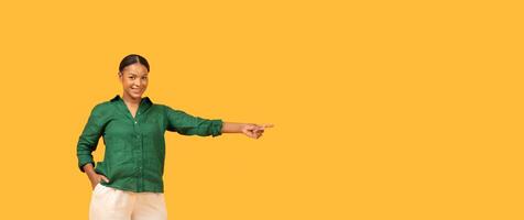 A black woman in a green shirt stands confidently, smiling and pointing to copy space. The vibrant yellow background adds energy to her cheerful expression, enhancing the lively atmosphere. photo