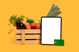A variety of fresh vegetables, including eggplant, carrots, and peppers, sit in a wooden crate alongside a tablet with blank screen and payment card on a vibrant yellow background. photo