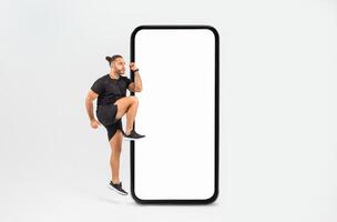 A man is playfully stepping up against a large blank smartphone display, showcasing an energetic pose. The indoor environment is simple and bright, emphasizing his enthusiasm. photo