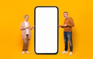 An elderly couple stands side by side, smiling as they point at a large blank screen. They are dressed casually, with a bright yellow backdrop enhancing the lively atmosphere of the moment. photo