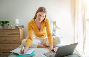 A teen girl sits cross-legged on her bed, focused on her laptop while jotting notes in a notebook. Sunlight streams through the window, creating a warm atmosphere in the room. photo