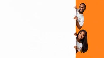 Intresting Offer. Happy Casual Black Couple Peeping Out The Side Of White Advertisement Board For Text Or Design. Smiling Cheerful People Holding Billboard, Looking At Camera, Standing On Yellow Wall photo
