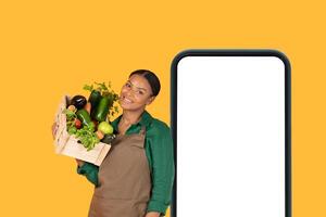 A black woman stands smiling while holding a wooden basket overflowing with fresh vegetables. She is positioned next to a large smartphone with a blank screen, bright yellow background. photo