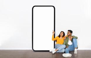 A young Indian couple sits casually on the floor, smiling and sharing a moment together near a large blank phone display. The setting is relaxed, with a simple background emphasizing their joy. photo