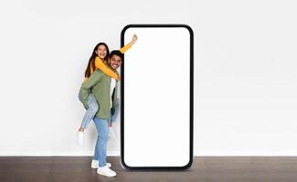 A cheerful Indian couple enjoys a playful moment in a bright space, with one partner giving a piggyback ride while standing next to an oversized smartphone photo