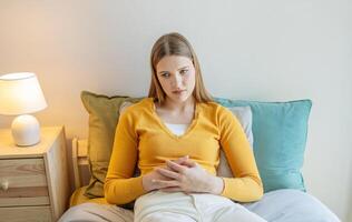 A teen girl is sitting on her bed with a worried expression, wearing a yellow sweater. The soft lighting and decor create a calm yet introspective atmosphere as she thinks quietly. photo