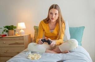 A teen girl sits cross-legged on her bed, focused on a game controller in her hands. A bowl of popcorn sits beside her, providing a cozy atmosphere in her bright room. photo
