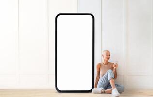 A woman in casual attire relaxes on the floor while using a smartphone. The space features a simple, clean design with a large blank smartphone placeholder beside her. photo
