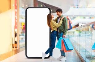 A young couple stands in a bustling mall, smiling at each other while holding colorful shopping bags. They are surrounded by retail displays and shoppers, creating a lively atmosphere. photo