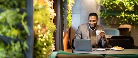 Adult black man holding white mug, smiling and looking at open laptop computer on table at cafe, copyspace photo