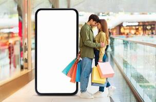 A young couple shares a moment of affection while holding colorful shopping bags. They are positioned beside a large blank smartphone display inside a modern mall setting. photo