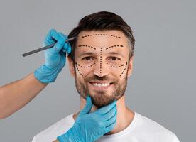 Surgeon hands making marks on smiling man face. Aesthetic Cosmetology, Plastic Surgery concept. Plastic surgeon applying marks with pencil on handsome man face before surgery, grey studio background photo