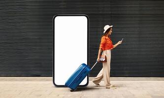 A woman walks with a blue suitcase while checking her smartphone, standing next to a large blank display. This urban setting suggests a busy travel hub during the day. photo