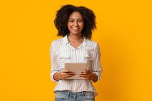 Millennial Afro Woman With Digital Tablet In Hands Smiling At Camera While Posing Over Yellow Background In Studio, Free Space photo
