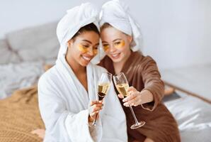 Happy multiethnic girlfriends with eye patches on toasting with champagne while having skincare procedures at home photo