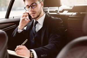 Always In Touch Concept. Young manager in glasses writing in notebook talking on smart phone sitting in back seat of car photo