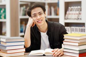 Education Concept. Cheerful asian boy sitting in library with open book, doing research, looking at camera, copyspace photo