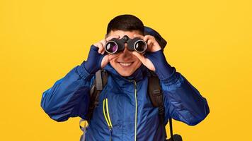 Portrait of smiling young guy hicker using modern binoculars, orange background photo
