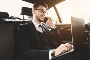 Always In Touch. Confident chief executive in suit using personal computer and making phone call in taxi, copy space, flare light photo