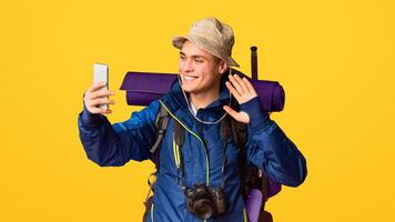 Smiling hiker in jacket with big backpack taking selfie on smartphone over orange studio background photo