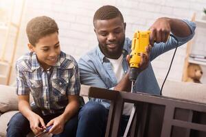 Black father teaching his son how to use drill, perforating chair in living room at home, sharing experience photo