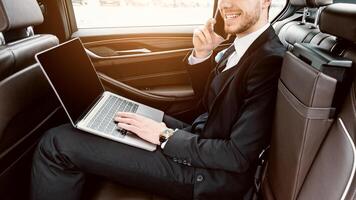 Always Available. Smiling male ceo in suit using laptop with blank screen talking on phone in car, copy space, side view photo
