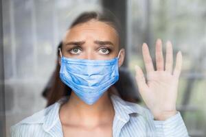 Social Distancing. Depressed Girl Wearing Face Mask Looking Out Of Window Standing At Home During Coronavirus Self-Isolation photo