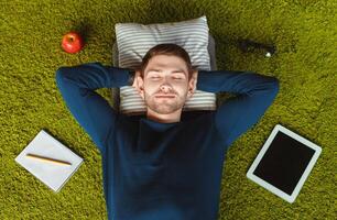 Happy University Student Guy Resting Lying On Green Grass Carpet Among His Belongings Tablet, Notebook. Above View photo