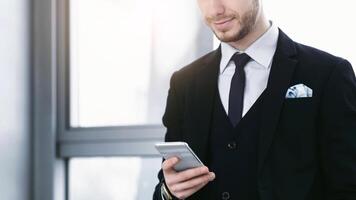 Good News From Colleague. Smiling man holding and looking at phone standing near window at his working place in office photo