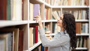 Young Librarian. Smiling mexican girl taking book from library bookshelf, searching and choosing manual, copyspace photo