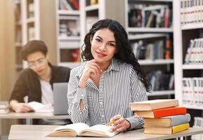 Learning And Educational Concept. Cheerful latin girl sitting at desk in public library, looking at camera, free space photo