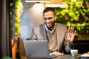 Coffee Break. Happy afro businessman in earbuds making call to family using laptop at cafe, waving hand, copyspace photo