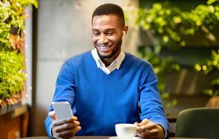 Technology Concept. Front view of happy afro student in headphone using smartphone sitting at modern cafe photo