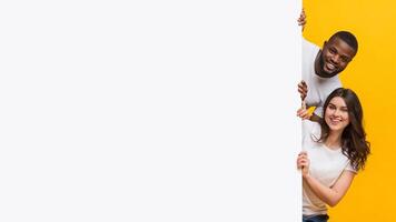 Joyful interracial couple peeking from behind white advertisement board with free space for your text, yellow studio background photo