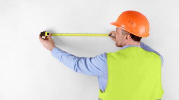 Professional Builder Measuring Wall With Tape-Measure Tool Standing Back To Camera Over White Studio Background, Wearing Orange Uniform. Construction Regulations, Building House. Panorama photo