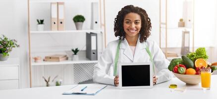 Smiling black lady nutritionist demonstrating digital tablet with empty screen, clinic interior, applications for dieting photo