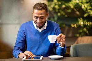 Technology Concept. Front view of happy black student sitting at modern cafe, drinking coffee and using tablet photo