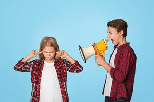 Misunderstanding Between Siblings. Little girl closing ears with fingers while her elder brother shouting to megaphone, blue background photo