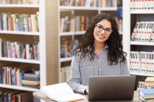 Doing Research. Front view of happy latina woman in specs using laptop computer in public library, smiling at camera photo