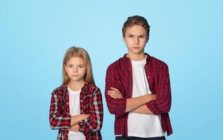 Siblings Conflicts. Portrait Of Frowning Little Brother And Sister Standing With Folded Arms And Looking At Camera, Blue Background With Empty Space photo