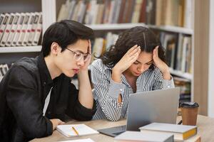 Two frustrated multicultural students sitting at campus library, doing research, trying to focus, copy space photo