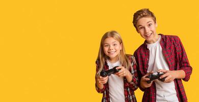 Kids Leisure. Brother And Sister Holding Joysticks, Playing Games Together, Posing On Yellow Background With Free Space photo