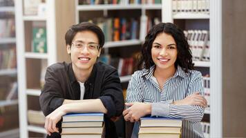 Front view of two smiling multicultural students leaning on pile of books and looking at camera sitting in modern library photo
